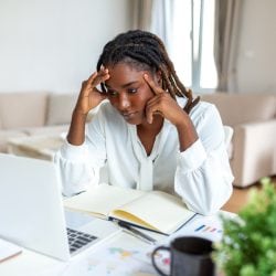 A young woman sits at a table comparing her laptop screen with a notebook.
